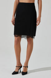 A woman is wearing a high-waisted black skirt with a lace hem peeking out from underneath. The skirt has a sleek, fitted style and the lace adds a decorative touch. She is also wearing black high-heeled shoes with ankle straps and gold rings on her fingers.